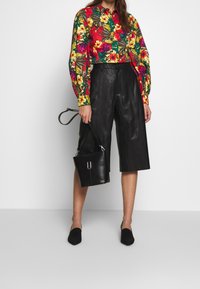 Chemise à boutons imprimé floral en rouge, vert et jaune, associée à des culottes en cuir noir taille haute et des chaussures plates noires. Sac à main noir inclus.