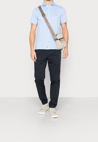 Camisa de manga corta azul claro con botones, pantalones azul marino oscuro y zapatillas deportivas beige claro. Bolso gris cruzado con cremallera y detalles de marca.