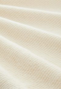 Texturerad crème-färgad tyg med ett subtilt ribbat mönster, som har mjuka, släta veck som visar dess lätta, vävda struktur.