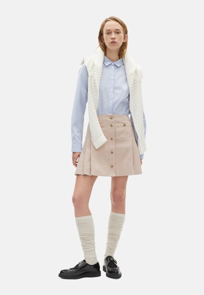 Lichtblauw gestreept overhemd met knopen, beige geplooide rok met gouden knopen, crème kniekousen en zwarte schoenen. Gebreide trui over de schouders gedrapeerd.