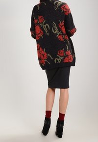 Cardigan de punto en color oscuro con patrones florales rojos, corte oversize y dobladillo acanalado. Combinado con una falda acanalada oscura y botas negras hasta el tobillo.