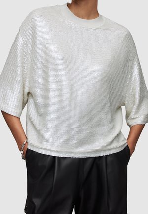Mannequin portant une chemise blanche ample à manches courtes en sequins assortie à un pantalon en cuir noir taille haute et un bracelet en argent.