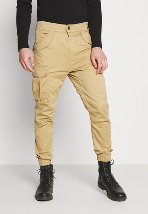 Khaki cargo pants with a button closure, side pockets, and elastic cuffs. Fabric has a smooth texture, paired with black lace-up boots.