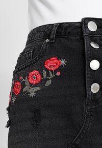 Svarta denimjeans med röd blomstermönstring, slitna detaljer och silverfärgad metall på knäppen.