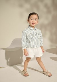 Enfant portant une chemise en denim rayée avec deux poches avant, un short blanc et des sandales beiges avec des accents de motif léopard.