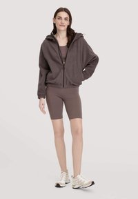 Braune Fleece-Zip-Jacke mit Kapuze, gerippten Bündchen und Saum; kombiniert mit einem passenden braunen eng anliegenden Oberteil und Fahrradshorts. Weiße Sportschuhe.