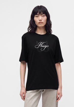 Frau mit schwarzen schulterlangen Haaren, die ein schwarzes Hugo-T-Shirt und hellbeige Hosen trägt, steht vor weißem Hintergrund.