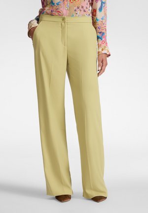 Vrouw draagt lichtgele wijd uitlopende broek met steekzakken en een kleurrijk gebloemde blouse met lange mouwen, gecombineerd met bruine puntige schoenen.