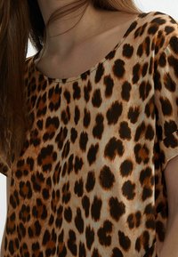 Primer plano de una mujer que lleva una blusa de manga corta con un patrón de leopardo marrón y negro y cabello largo castaño.