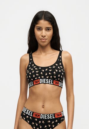 Frau trägt schwarzen floralen Sport-BH und passende Unterwäsche mit markanten Diesel-Logo-Bändern auf einem schlichten hellen Hintergrund.
