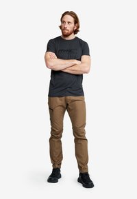 Mörkgrå T-shirt med en logotyp, ihop med ljusbruna byxor med dragkedjeficka och elastiska muddar. Svarta skor fulländar outfiten.