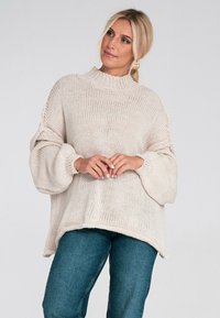 Beżowy, oversize sweter z dzianiny o wysokim dekolcie, prążkowanej teksturze i bufiastych rękawach, w połączeniu z ciemnymi dżinsami.