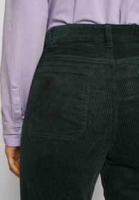 Personne portant un pantalon en velours côtelé vert foncé et une chemise à manches longues violet clair avec des poignets boutonnés, montrée de la taille jusqu'en bas.