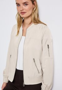 Beżowa kurtka bomber z metalowymi zamkami błyskawicznymi, okrągłym kołnierzem i ściągaczami. Posiada dwie boczne kieszenie oraz kieszeń na rękawie zapinaną na zamek.