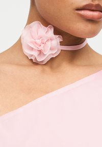 Κοντινό πλάνο ενός ατόμου που φοράει ένα διάφανο ροζ κολιέ με λουλούδια και ένα ανοιχτό ροζ τοπ χωρίς ώμους, με φόντο έναν απλό χώρο.