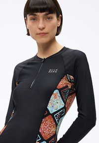 Kobieta o krótkich ciemnych włosach nosi czarną bluzę na zamek z długim rękawem, z kolorowymi geometrycznymi kwiatowymi panelami oraz logo "Billabong" na piersi.