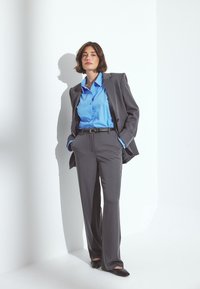 Un costume gris ajusté avec un pantalon à jambes larges et une veste assortie. En dessous, une chemise à boutons bleu clair, complétée par une ceinture noire et des chaussures.