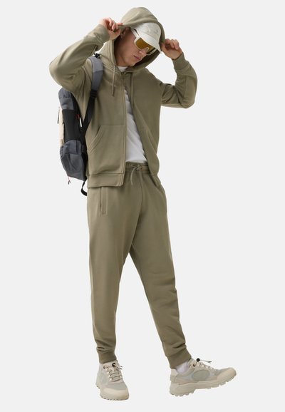 Olivegrüner Hoodie mit Reißverschluss und Taschen, kombiniert mit passenden Jogginghosen. Das Outfit wird durch eine weiße Cap und Sonnenbrille ergänzt.