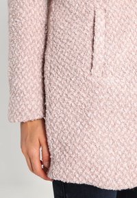 Lichtroze textuurjas van een zacht, pluizig materiaal. Beschikt over één zak en lange mouwen, met een rechte zoom aan de onderkant.