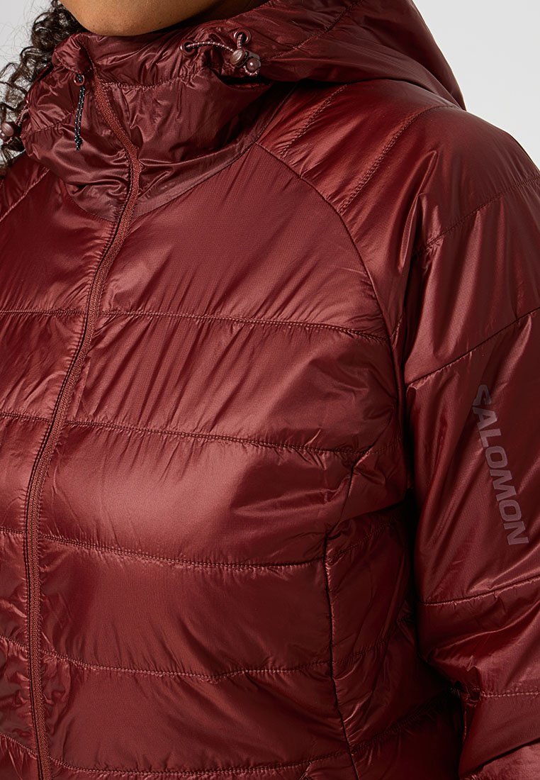 Chaqueta aislante color marrón con acabado brillante, cierre frontal con cremallera y capucha con cordón. Presenta costuras horizontales y el logo "Salomon" en la manga.