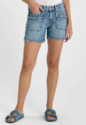 Vrouw die lichtblauwe denimshorts draagt met grote voorzakken en blauwe sandaal met gesp, staand tegen een effen achtergrond.