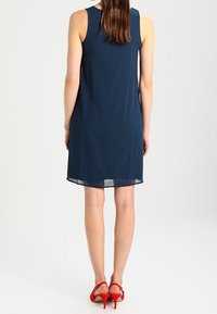 Laulullinen, laivastonsininen ilman hihoja oleva mekko, jossa on rento leikkaus ja läpikuultava helma. Yhdistettynä punaisiin niittikorkoihin, mikä korostaa sileää kangasrakennetta.