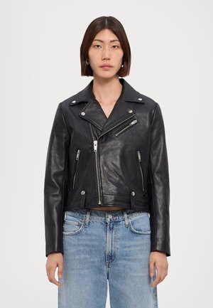 Vrouw draagt een zwarte leren bikerjas met zilveren ritsen en lichtblauwe spijkerbroek, staand tegen een effen witte achtergrond.