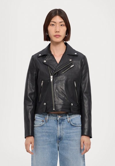 Kvinde iført en sort læderbikerjakke med sølv lynlåse og lyseblå jeans, stående mod en ensfarvet hvid baggrund.