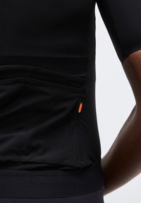 Κοντινή λήψη ενός ατόμου που φοράει μαύρο εφαρμοστό πουκάμισο με μικρό πορτοκαλί φερμουάρ σε μια πίσω τσέπη, δείχνοντας κοντά μανίκια και μέρος του χεριού.