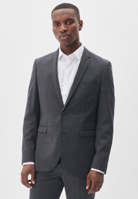 Traje gris con un acabado texturizado, chaqueta de un solo pecho, solapas en pico y dos bolsillos frontales. Lleva una camisa blanca con cuello debajo.