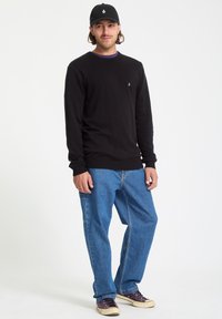 Czarny sweter z długim rękawem i ściągaczami przy mankietach, niebieskie dżinsowe spodnie oraz czapka w kolorze czarnym z małym logo z przodu. Sneakersy z purpurowymi akcentami.