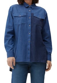 Kvinde iført tofarvet blå denim skjorte med knapper og brystlommer samt matchende blå jeans, der står foran en hvid baggrund.