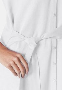 A hand adjusts a tied belt on a plain white shirt, featuring buttons down the front and a smooth, textured fabric.