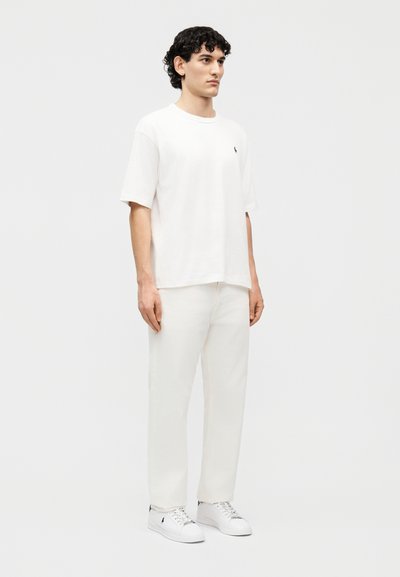 Άνδρας με σγουρά σκούρα μαλλιά στέκεται φορώντας λευκό φαρδύ μπλουζάκι, λευκό παντελόνι και λευκά αθλητικά παπούτσια μπροστά σε απλό ανοιχτό φόντο.