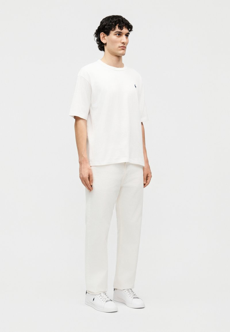 Man met krullend donker haar staat, gekleed in een oversized wit T-shirt, witte broek en witte sneakers, tegen een eenvoudige lichte achtergrond.