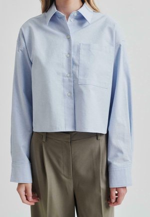 Frau trägt ein hellblaues, kurz geschnittenes Hemd mit Knöpfen, einer Brusttasche und langen Ärmeln, kombiniert mit hoch taillierten olivgrünen Hosen.