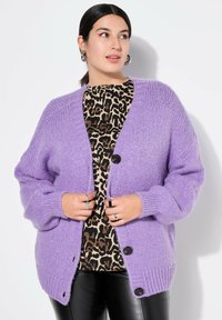 Lavendelfarbene Strickjacke im Oversized-Schnitt mit Knopfverschluss und gerippten Bündchen. Getragen über einem leo-musternden Oberteil und schwarzen Kunstlederhosen.