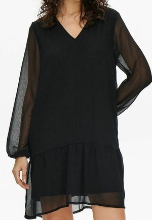 Kobieta ubrana w czarną sukienkę z dekoltem w serek, przejrzystymi długimi rękawami i wielowarstwowym, przejrzystym dołem sięgającym powyżej kolan.