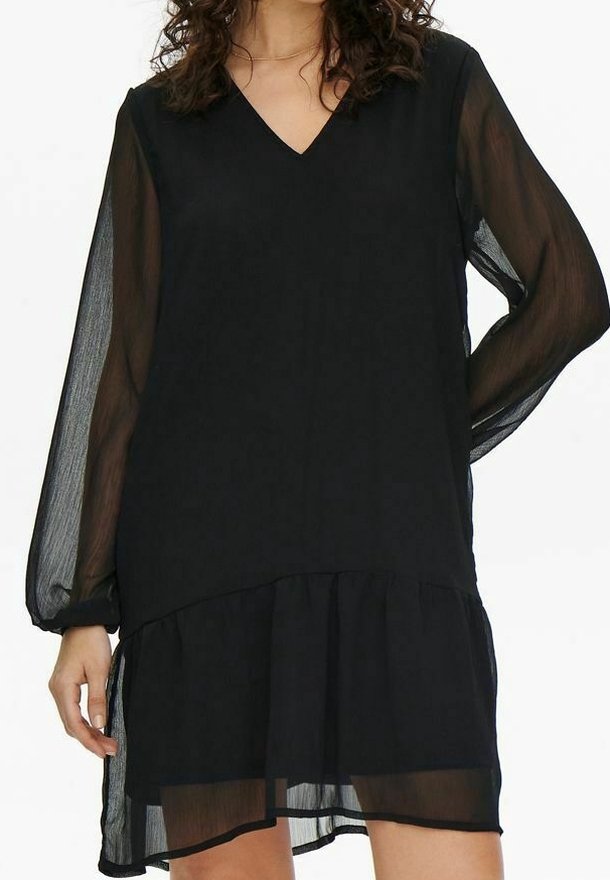 Femme portant une robe noire à col en V avec des manches longues transparentes et un bas superposé transparent au-dessus des genoux.