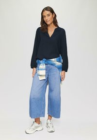 Femme portant un chemisier noir à manches longues, un jean bleu clair à taille large, des baskets blanches, et un pull en tricot bleu noué autour de la taille, se tenant debout.