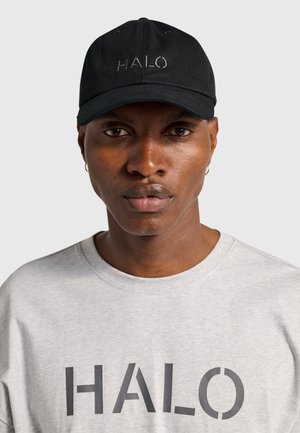Schwarze Mütze mit dem Schriftzug „HALO“, getragen von einer Person in hellgrauem Hemd, das ebenfalls den Schriftzug „HALO“ zeigt, vor schlichtem hellem Hintergrund.