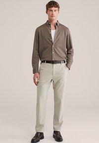 Jeune homme portant une chemise marron à boutons par-dessus un t-shirt blanc, un pantalon beige avec une ceinture noire, et des mocassins marron foncé, debout devant un fond uni.