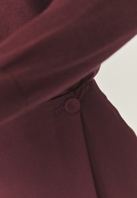 Bordový oděv s hladkou texturou, akcentem na knoflík a přizpůsobeným designem s detailním skladem. Zaměření na látku a zpracování.