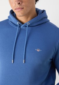 Prikaz osebe v modri kapucarji z zankami in majhnim logotipom GANT na prsih, svetla koža z vidnimi pegicami.