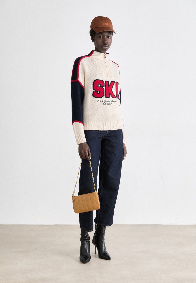 Trui met marineblauwe en crèmekleurige blokken, rode accenten en "SKI"-logo. Donkerdenim broek en zwarte hoge hakken. Houdt een gekwilt tan handtas vast.