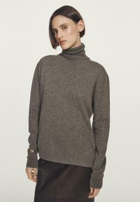 Frau mit kurzen braunen Haaren, die einen grauen Rollkragenpullover und einen dunklen Rock vor einem schlichten hellen Hintergrund trägt.