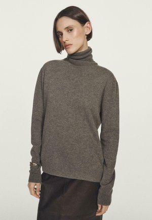Donna con capelli corti castani che indossa un maglione grigio a collo alto e una gonna scura su uno sfondo chiaro uniforme.