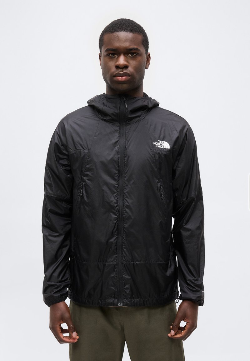 Άνδρας που φοράει μαύρο μπουφάν The North Face με φερμουάρ και κουκούλα, στέκεται μπροστά σε απλό λευκό φόντο.