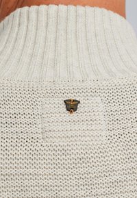 Witte gebreide trui met een geribde kraag, waaronder een klein zwart en gouden "PMF LEGEND" logo op de achterkant en een gedetailleerde textuur.