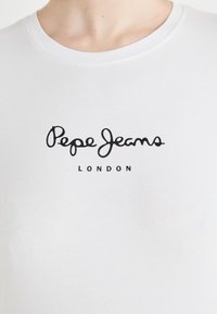 Valkoinen puuvillapaita, jossa on pyöreä pääntie. Paidassa on mustalla kirjoitettu "Pepe Jeans" ja sen alla pienemmällä suoralla fontilla "LONDON".
